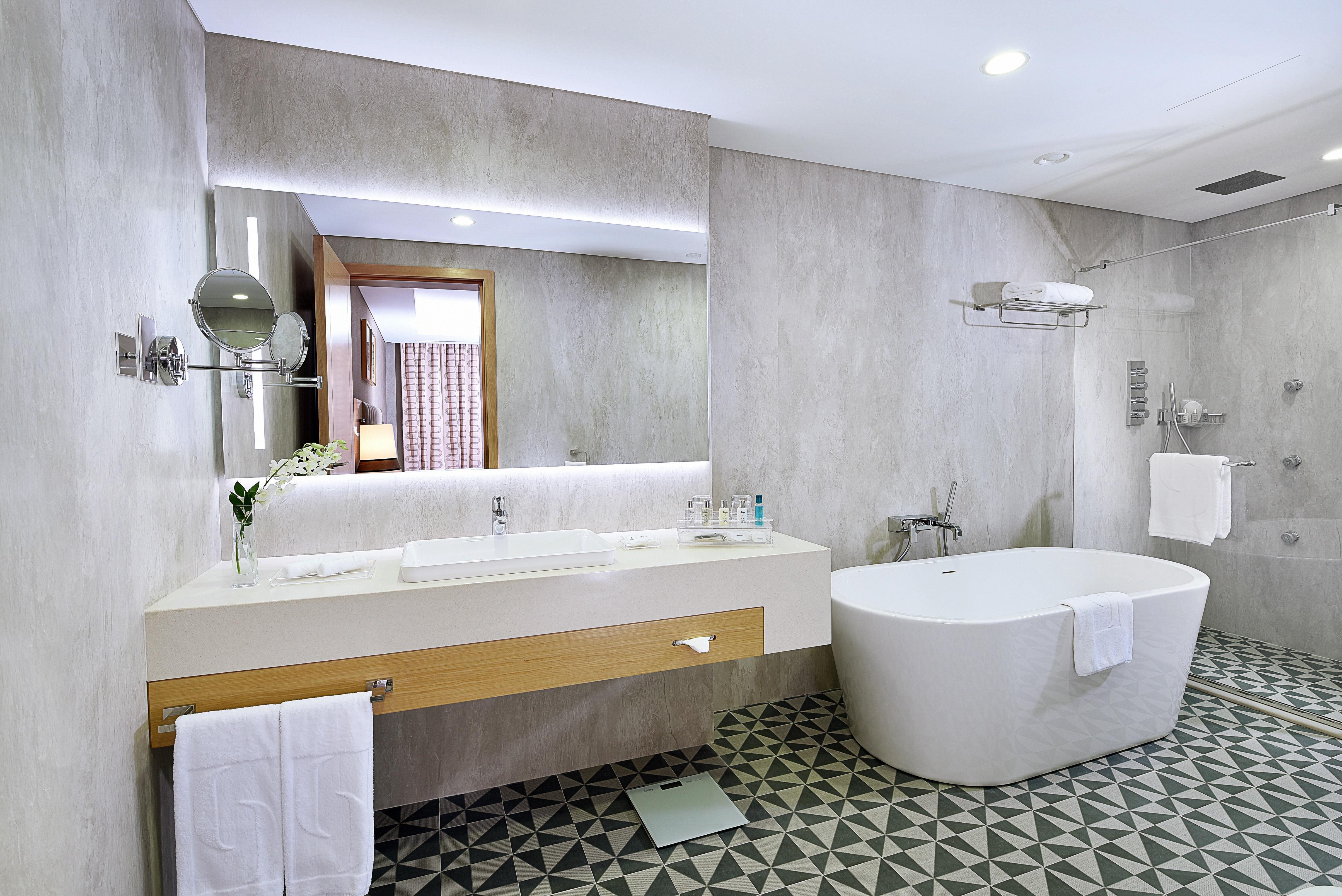 Grand Cosmopolitan Hotel Dubai Exterior foto The photo shows a modern bathroom featuring a sleek design. There is a large white freestanding bathtub on one side. The vanity area has a long countertop with a sink and a large mirror above it, illuminated by soft lighting. The walls are a light gr