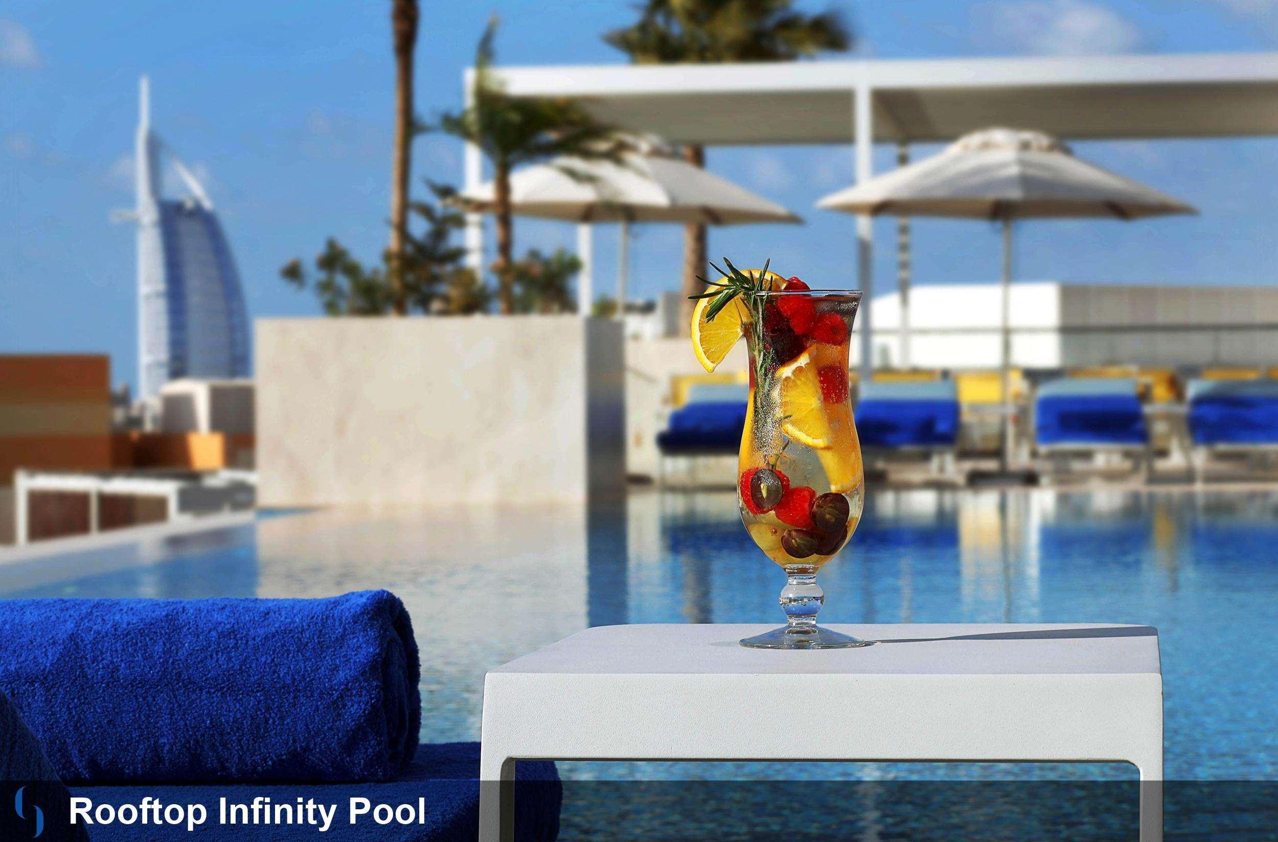 Grand Cosmopolitan Hotel Dubai Exterior foto The photo shows a rooftop infinity pool with a stunning view. In the foreground, there is a cocktail glass filled with a colorful drink garnished with fruits, including lemon and berries. The background features cozy loungers and umbrellas, suggestin