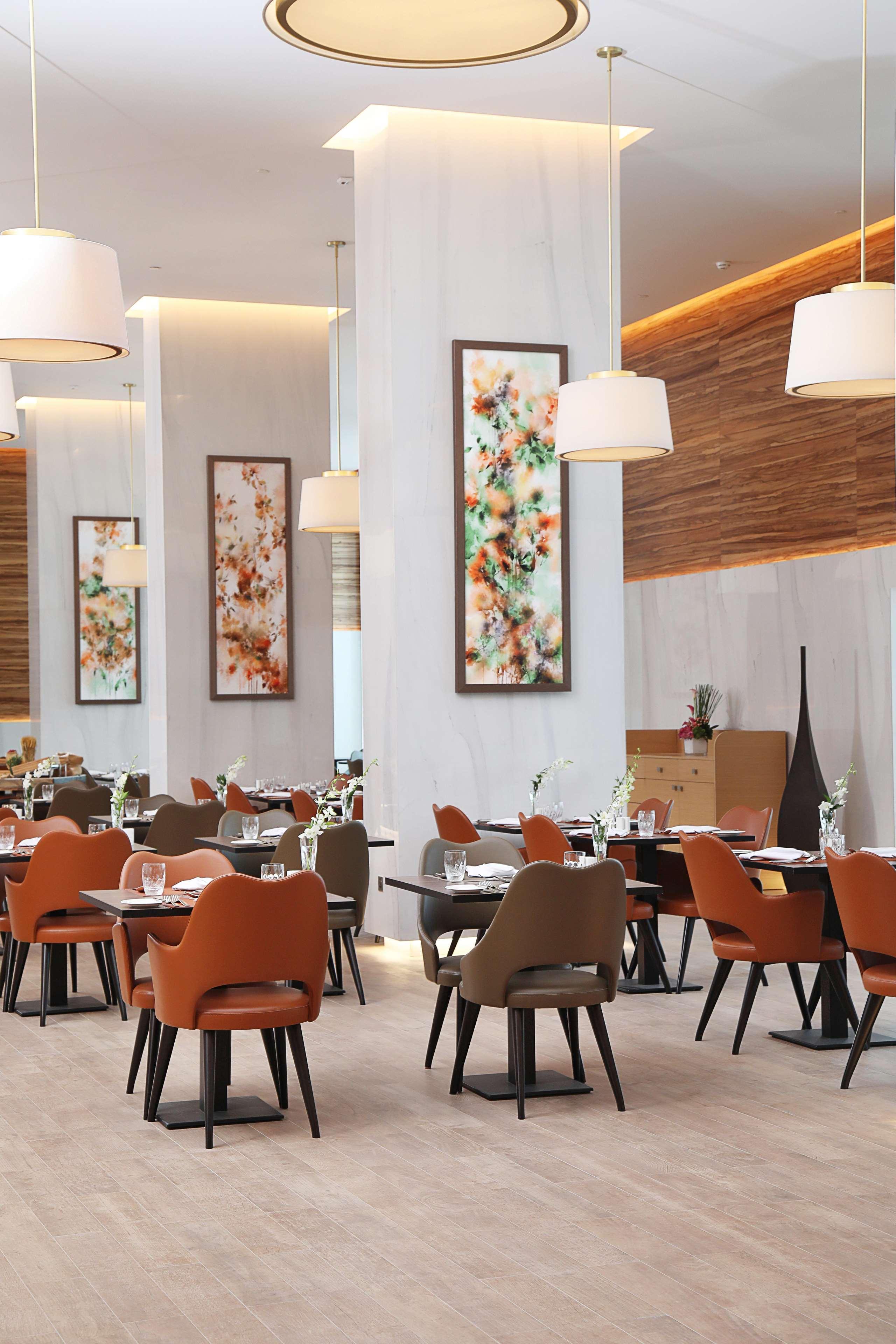 Grand Cosmopolitan Hotel Dubai Exterior foto The photo shows the interior of a modern restaurant. The space features elegantly set dining tables with glasses and cutlery, and the chairs have a mix of colors, including orange and neutral tones. There are large pieces of artwork on the walls, lik
