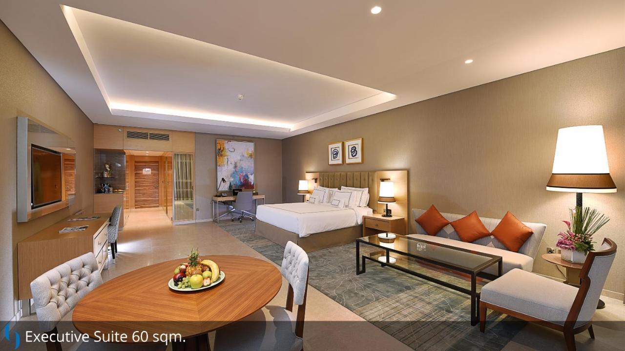 Grand Cosmopolitan Hotel Dubai Exterior foto The image shows a spacious hotel suite. It features a king-sized bed with a modern design, accompanied by decorative pillows. To one side, there is a seating area with a sofa and a glass coffee table. A dining table with chairs is also visible, posit