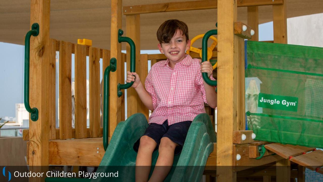 Grand Cosmopolitan Hotel Dubai Exterior foto The image shows a young boy sitting on a slide in a children's playground. He is wearing a pink checkered shirt and dark shorts. The playground equipment is made of wood and features green sliding elements. The boy has a joyful expression, suggesting