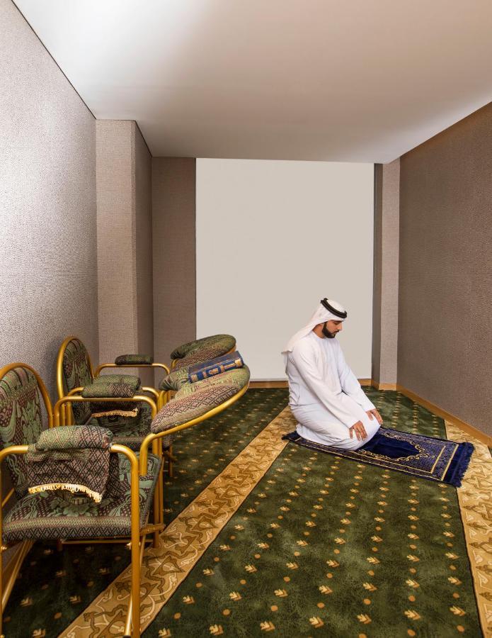 Grand Cosmopolitan Hotel Dubai Exterior foto The image shows a simple, serene indoor setting. In the center, a man wearing traditional attire is kneeling on a prayer rug, engaged in prayer. Surrounding him are several ornate chairs, and the floor is adorned with patterned carpets. The walls are